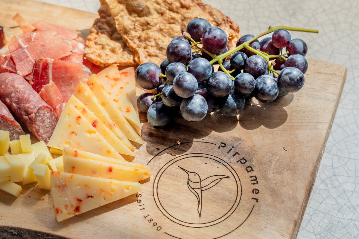 Speck, Salami, Käse, Schüttelbrot und Trauben auf einem Holzbrett mit Pirpamer-Logo.